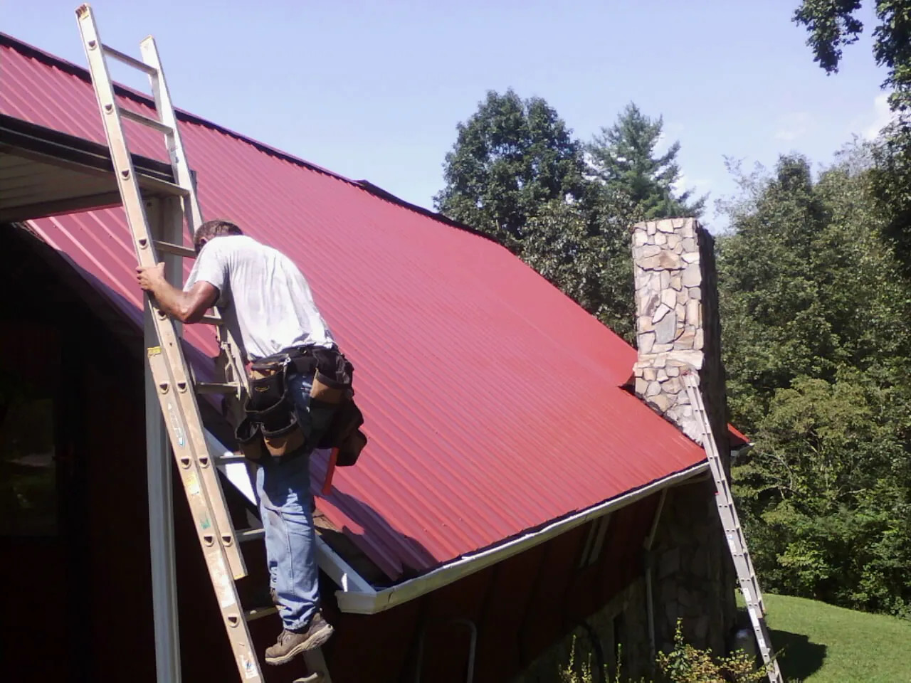 Roofer on a ladder beside a red metal roof and stone chimney — Soffit & Fascia Repair by Roofing Brothers in Eldorado